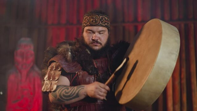 Asian man with tattooed arms conducts evil spirits placating with shamanic drum in ail closeup. Festival of Siberian local culture and traditions