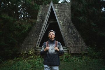 Stranger traveler man in ethnic sweater alone standing in front of old vintage style wooden hut in the dark forest. Scandinavian style moody aesthetic image