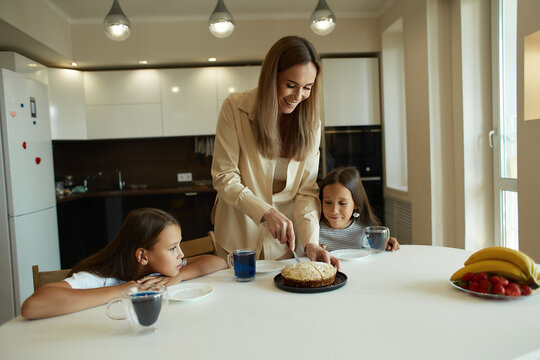 Charming attractive beautiful caucasian kind young smiling mother is drinking tea with her daughters, dressed in casual clothes, sitting in a bright kitchen, cutting pirok