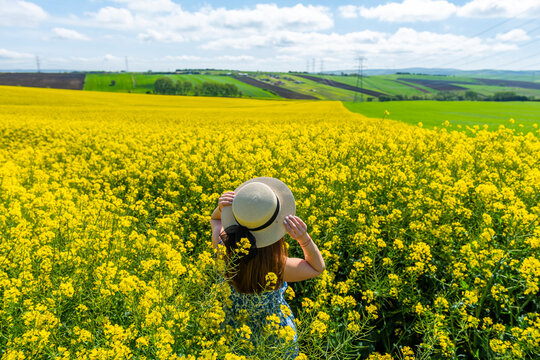 Beautiful girl with dress and hat in the Canola Fields. Blooming canola fields under a blue sky with clouds. Beautiful yellow flowers. Happy girl in nature.