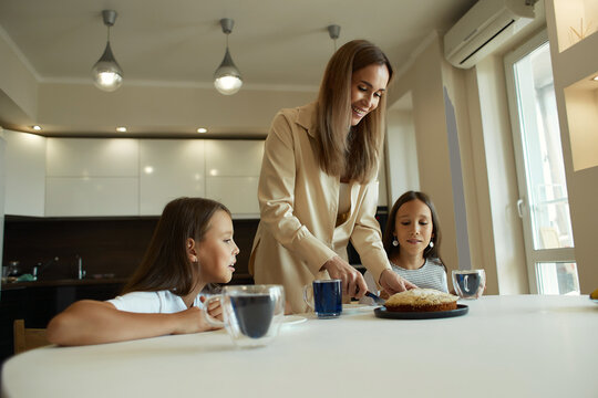 Charming attractive beautiful caucasian kind young smiling mother is drinking tea with her daughters, dressed in casual clothes, sitting in a bright kitchen, cutting pirok