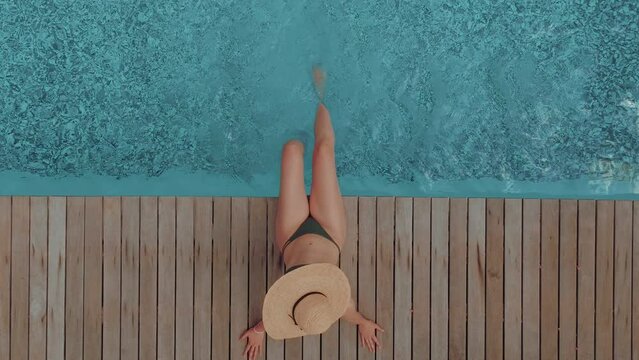 Young woman wearing a straw hat enjoying by the blue swimming pool. Relaxing vacation/holiday on a hot summer day. Escaping the town and busy lifestyle.