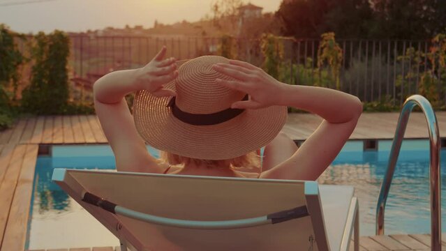 Young woman wearing a straw hat enjoying on swimming pool lounge, laying on a sun chair and looking at the beautiful sunset.  Relaxing vacation / holiday. Escaping the town and busy lifestyle. 
