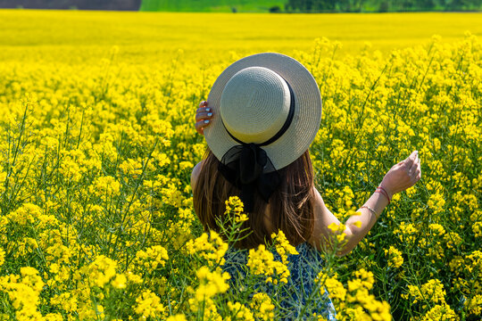 Beautiful girl with dress and hat in the Canola Fields. Blooming canola fields under a blue sky with clouds. Beautiful yellow flowers. Happy girl in nature.