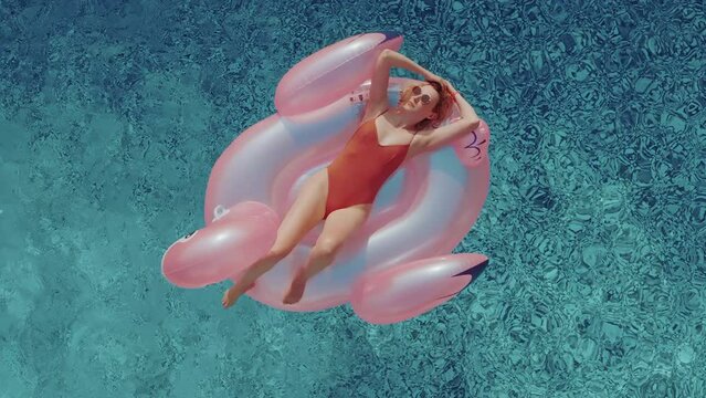 A young tourist woman with blonde hair is sunbathing on an inflatable pink flamingo in the middle of a swimming pool on a hot summer day. Female relaxing on a raft in the pool on a sunny day.