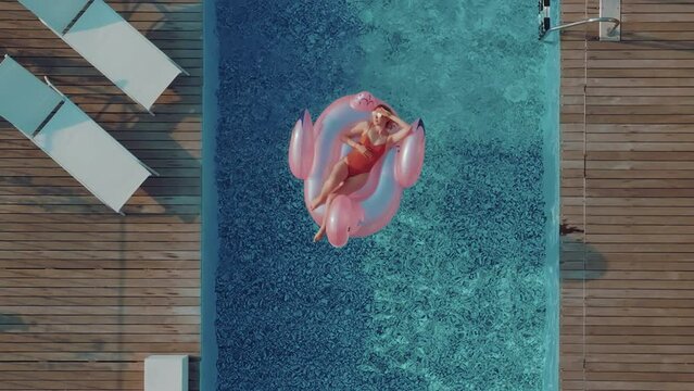 A young tourist woman with blonde hair is sunbathing on an inflatable pink flamingo in the middle of a swimming pool on a hot summer day. Girl relaxing sunbathing and enjoying travel holidays at a res