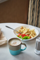 Cappuccino coffee in cup and plate with scrambled eggs and tomatoes. Breakfast on the table