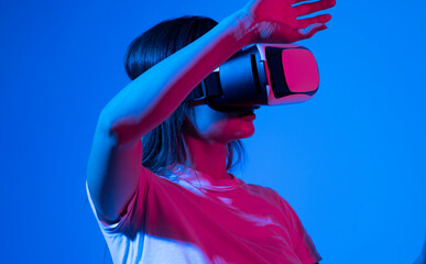 Excited brunette woman in white t-shirt getting experience using VR-headset glasses of virtual reality much gesticulating hands in neon light.