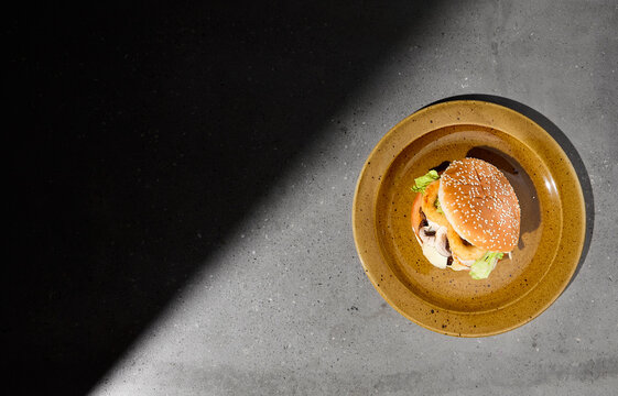American burger with onion rings on ceramic plate in dark minimal style. Beef burger on gray stone table with hard shadow. Fast food dark concept. Burger on gray concrete background. Simple food menu.