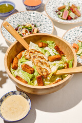 Classic caesar salad with grilled chicken and crouton in wooden bowl on white table. Caesar salad with sauce and wooden spoon. Italian lunch with caesar salad on sunny day. Summer menu.