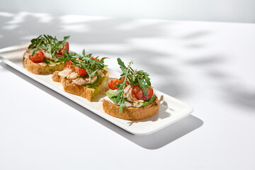 Aesthetic composition with shrimp bruschetta on white background with shadows from flowers. Italian bruschetta with shrimp, tomatoes and arugula on fine dining in summer. Elegant menu concept.