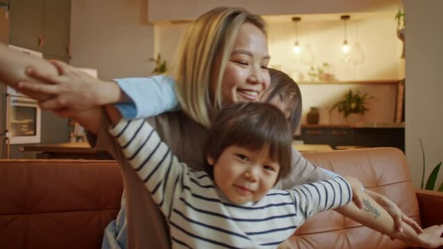 Happy Asian Family Loving Young Mum Playing Piggybacking Cute Little Funny Kids Sit on Couch At Home, Cheerful Mother Carrying Small Childs on Back Bonding Pretending Flying Having Fun Together