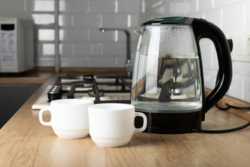 Modern electric transparent kettle with tea cups on a wooden table in the kitchen. Kettle for boiling water and making tea. Household appliances for making hot drinks. Place to copy. Place for text.