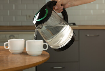 A man makes tea using boiling water from an electric kettle in the kitchen at home. It's time to have breakfast and drink tea. Modern electric kettle on a wooden table. Kettle for boiling water.