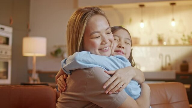 Happy Little Asian Cute Girl Hugging, Embracing Her Mom Enjoying, Bonding, and Laughing Together. Mother Parent Hugging her Kid Having a Tender and Lovely Moment in Living Room at Home