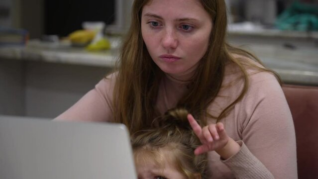 Busy young mother is trying to work remotely with her child at home. Yelling crying daughter interferes with mom work on laptop. Woman tries to distract her little daughter from hysterics
