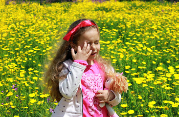 Fototapeta premium Curly little girl with a red bow in her hair. Girl with a dol on a green meadow among yellow flowers