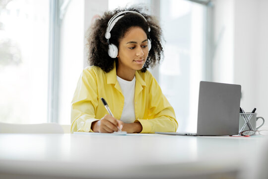 Portrait of a busy focused black woman in a wireless headset writing in a notepad sitting at a work desk with a personal computer in a modern office, profile side view