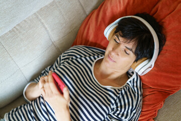 Woman consulting her smartphone while listening to music with headphones lying on the sofa.