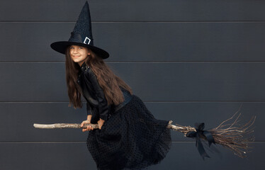 Portrait of cute teen girl in a Halloween witch costume with hat flies on a broom on a black wall...