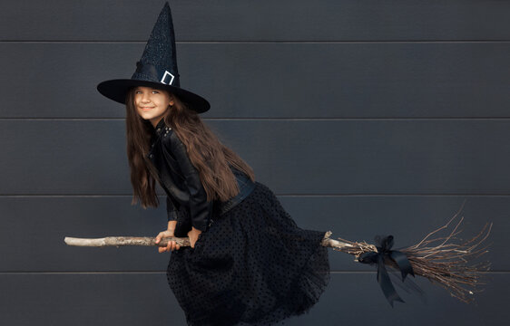 Portrait of cute teen girl in a Halloween witch costume with hat flies on a broom on a black wall background