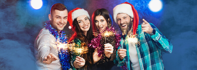 A group of young adult cheerful friends at a party wearing santa hats and tinsel with sparklers in...