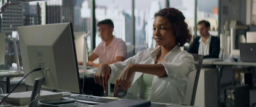 Bored African American female employee silly typing on a keyboard at her working desk in a large busy office, other workers in the background