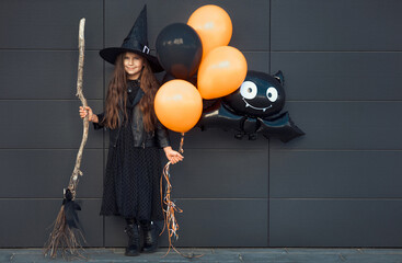 Full body little girl in black witch costume with hat and broom holding balloons with bat, standing on city street at the dark wall 