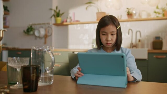 Curious Asian Preschool Kid Girl Using Digital Tablet Technology Device Sitting by Desk Alone. Small Child Hold Pad Computer Surfing Internet Play Game at Home. Children Tech Addiction Concept