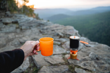 A hand holds a mug of tea against the backdrop of the sunset in the mountains, a hiker drinking a...