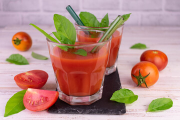 Two glasses of tomato juice and fresh basil leaves on a white background.
