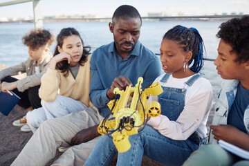 Portrait of young black girl holding robot model during engineering class outdoors with male...