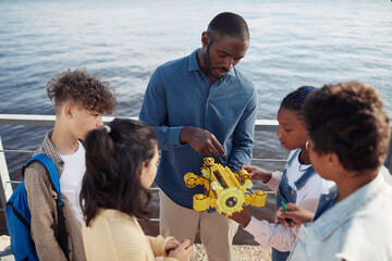 Portrait of black male teacher demonstrating robot model to group of children standing in circle...