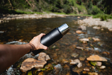 The guy holds a thermos in his hand against the background of the river, his hand holds a thermo bottle. trekking on a mountain river alone, hiking in extreme conditions. - obrazy, fototapety, plakaty