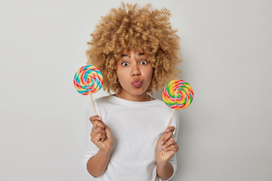 Horizontal shot of lovely curly haired woman keeps lips rounded holds two big colorful lollipops has sweet tooth dressed in casual jumper isolated over grey background. Unhealthy eating concept