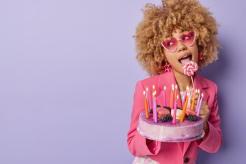 Horizontal shot of stylish beautiful woman with curly hair licks sweet candy holds delicious sweet cake with burning candles going to celebrate birthday looks aside isolated over purple background