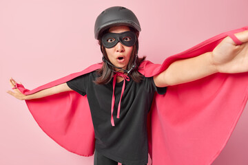 Shocked adult woman superhero stretches arms prepares for flight stares amazed with opened mouth dressed in costume with cloak poses against pink background. Ready to save world and help you