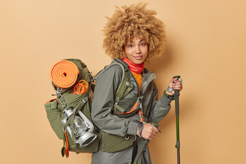 Horizontal shot of active curly female hiker poses with scandinavian poles climbs in mountains has camping trip carries heavy rucksack full of equipment for picnic isolated over beige background