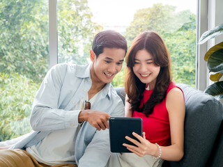 Happy young beautiful Asian woman and handsome man smile together while looking at tablet screen on grey sofa near the glass window at white living room. Couple lover using digital tablet together.