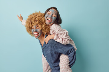 Positive young women friends have fun together give piggyback ride wear slumber suits apply beauty mud mask undergo skin care procedures isolated against blue background. Friendship concept.