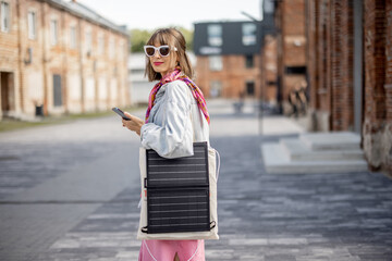 Young stylish woman walks with smart phone and charge it from portable solar panel hanging on bag....