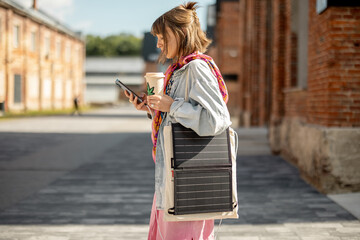 Young stylish woman walks with coffee and smart phone charging it from portable solar panel hanging...