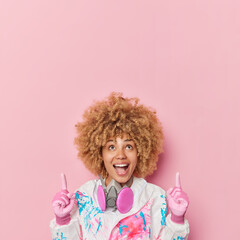 Obraz na płótnie Canvas Glad surprised woman wears biohazard chemical protective suit and respirator indicates above on blank space demonstrates advertisement isolated over pink background. Female physician in uniform