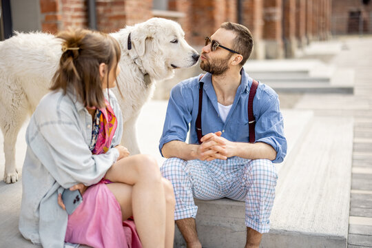 Young stylish couple sit together and have fun with their white dog on a street. Young hipsters hang out together near office outdoors