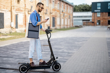 Fototapeta na wymiar Stylish guy drives electric scooter and charges phone with solar panel on the move on a street at office district. Sustainable business lifestyle and green energy concept