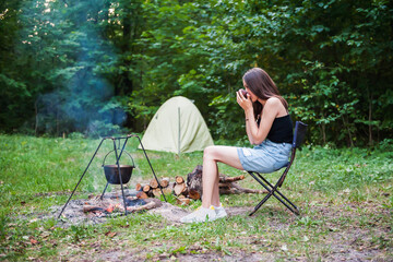 A girl drinks tea by the fire against the backdrop of a forest and a tent in summer