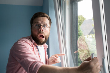 Shocked man with a strange expression on face stands near an open window and points finger into the...
