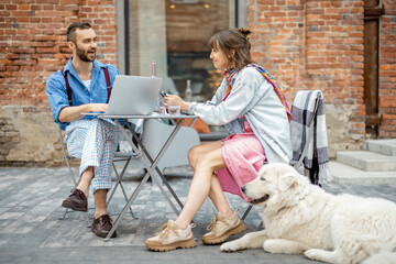 Stylish couple of colleagues have a conversation while sitting with dog at cafe terrace in modern...
