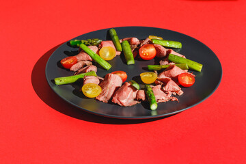 Roast beef with tomatoes and asparagus, dish on red background. Hard light, deep shadow