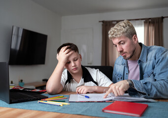 Schoolboy looking exhausted while father tries to help him with homework.